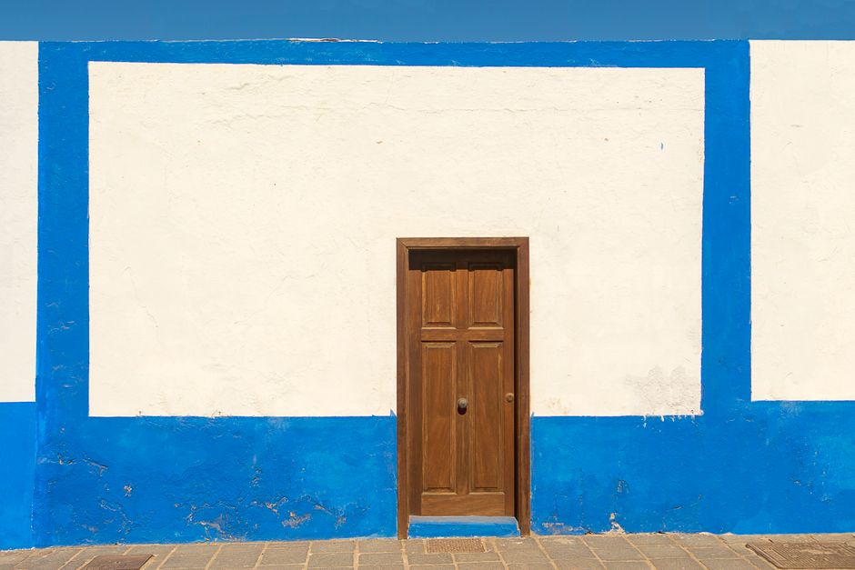 A rustic wooden door set in a striking blue and white wall under a clear sky.